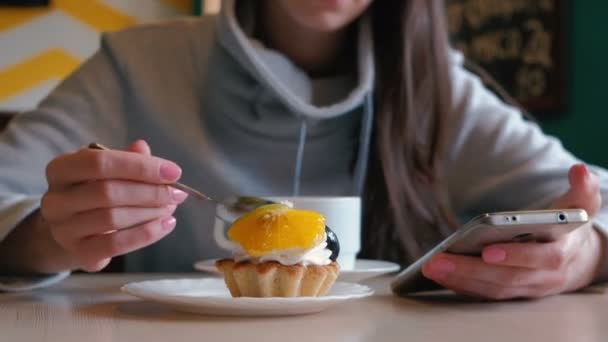 Unrecognizable woman eats a cake with a spoon and looks at the phone screen. Close up. — Stock Video