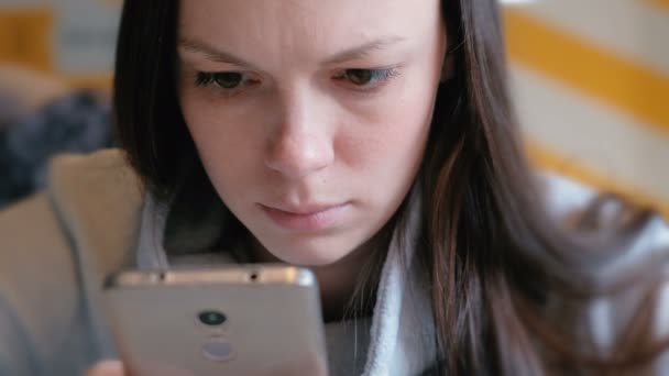 Młoda kobieta poważnie, czytając coś na jej telefon komórkowy siedząc w kawiarni. Zbliżenie twarzy. — Wideo stockowe