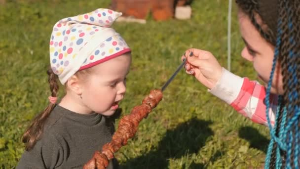 Maman nourrir sa jolie petite fille avec viande shashlik barbecue sur une brochette dans le jardin . — Video