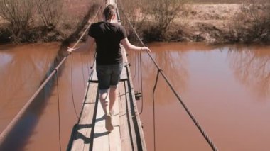 Genç adam ahşap Asma köprüde Nehri üzerinde yürüyor. Arkadan görünüm.