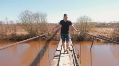 Genç adam ahşap Asma köprüde Nehri üzerinde yürüyor.