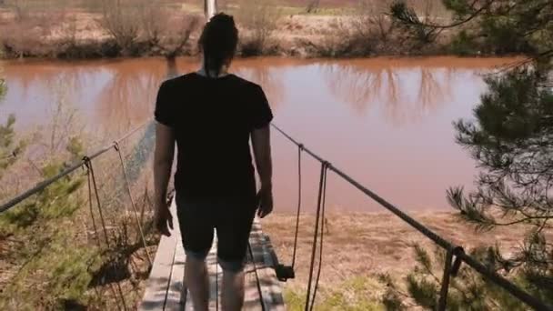 O jovem está andando sobre uma ponte de madeira suspensa sobre o rio. Visão traseira . — Vídeo de Stock