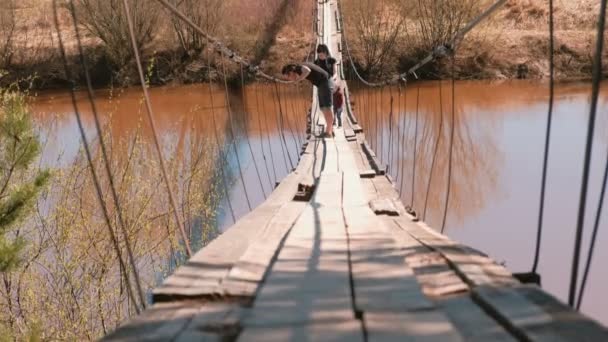 Jeune famille maman, papa et fille marchent sur le vieux pont suspendu au-dessus de la rivière . — Video