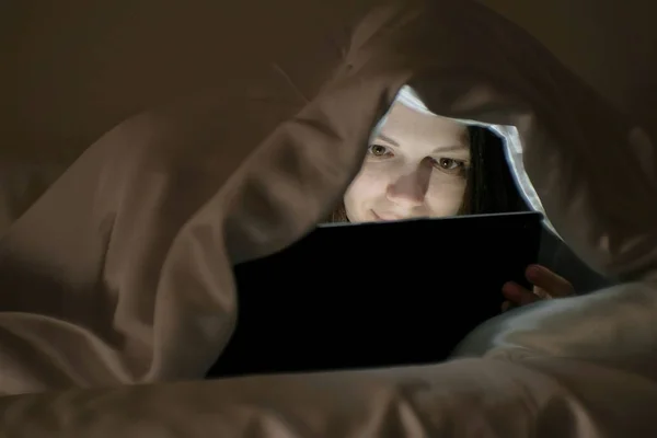 Νεαρή γυναίκα βλέποντας μια αστεία ταινία σε δισκίο κάτω από την κουβέρτα. — Φωτογραφία Αρχείου