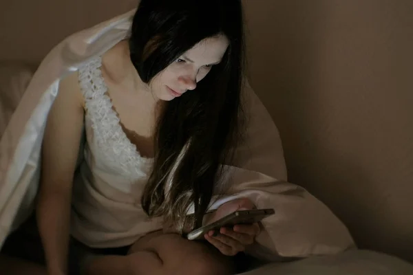 Joven hermosa mujer morena mira los mensajes en su teléfono móvil en la cama antes de dormir . — Foto de Stock
