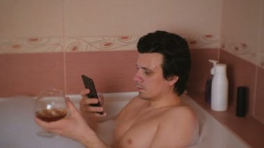 Genç adam bir banyo köpüğü, türleri haber üstünde hareket eden telefon ve içki viski ve gülümsüyor ile alır.