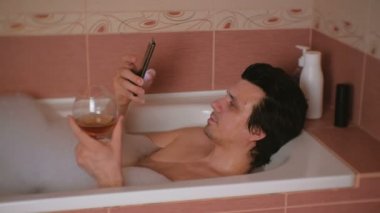 Genç adam bir banyo köpük, cep telefonu, içki viski ve kameraya bakarak gülümser Internet tarama alır.
