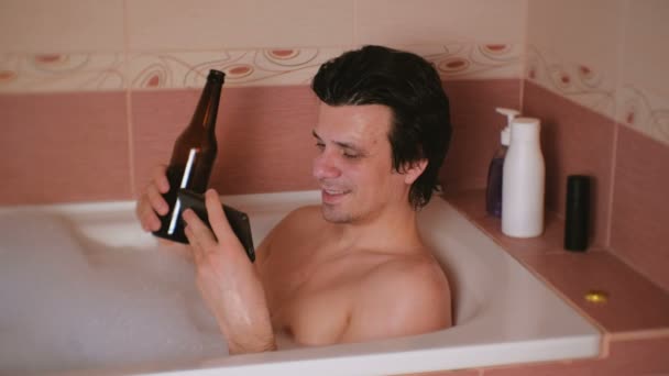 Joven toma un baño con espuma, mira videos divertidos en el teléfono móvil, bebe cerveza de botella . — Vídeo de stock