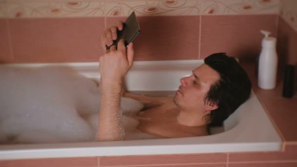 Young man takes a bath, watches the video on the tablet seriosly. — Stock Video