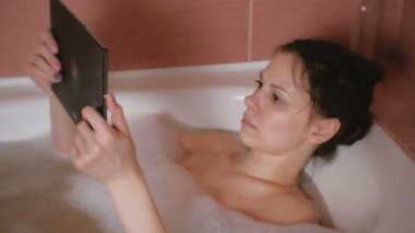 Genç güzel esmer kadın banyo alır ve seriosly tablet video izler.