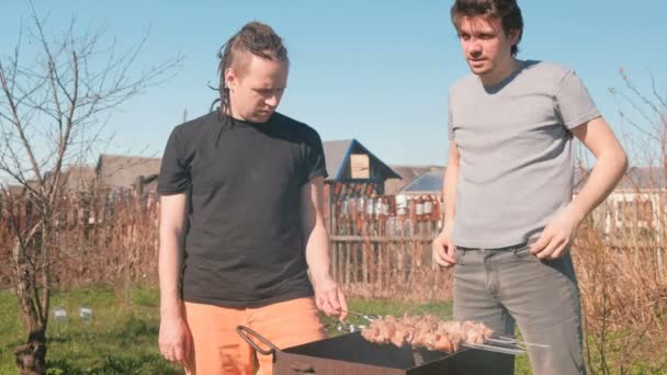 Dos amigos hombres cocinan carne shashlik en la parte superior de la parrilla de carbón en el patio trasero. Hablando y sonriendo juntos . — Vídeos de Stock