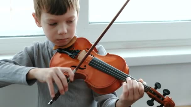 Toca violino. Um rapaz de sete anos a tocar violino perto de uma janela. Vista frontal . — Vídeo de Stock