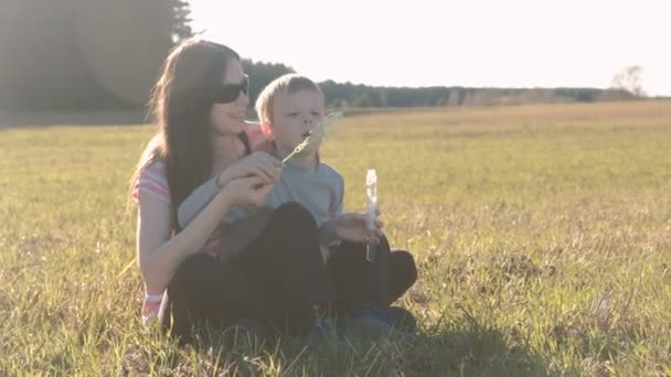 Moeder en zoon waait een zeep bubbels zittend op het gras, familie weekend in park. — Stockvideo