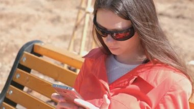 Güneş gözlüklü genç esmer kız parkta bankta oturan telefonda bir video izliyor.