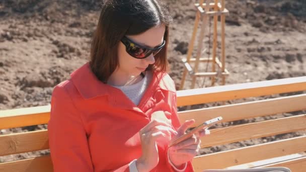 Jonge brunette meisje in zonnebril schrijft en een signaal aan de telefoon zitten in het Park op de Bank. — Stockvideo