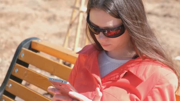 Молода брюнетка в сонцезахисних окулярах дивиться відео на телефон, сидячи в парку на лавці . — стокове відео