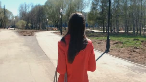 Una joven con abrigo rojo camina por el parque de primavera. Vista trasera . — Vídeo de stock
