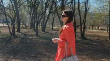 Genç kadın esmer kırmızı paltolu güneş gözlüğü bahar Park yürür.