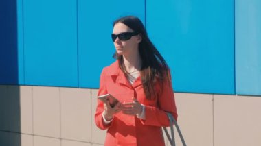 Genç kadın esmer güneş gözlüğü kırmızı paltolu biri için bekler ve onun telefon denetler manifatura.