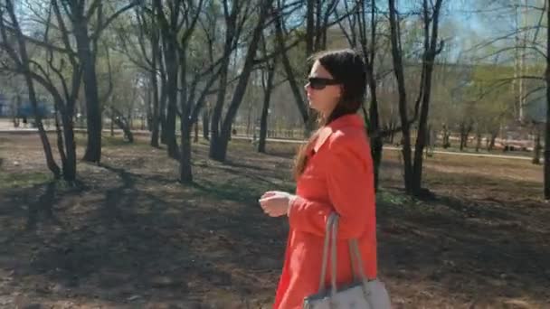 Jeune femme brune en lunettes de soleil en manteau rouge marche au printemps Park . — Video