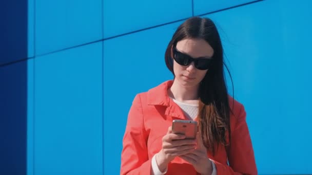 Jeune femme brune en lunettes de soleil en manteau rouge attend quelqu'un et vérifie son téléphone, textos . — Video