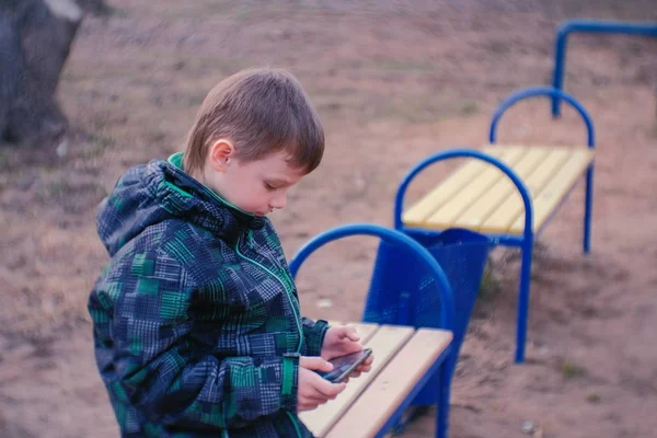 Pojke spelar ett spel på sin mobiltelefon som sitter i parken på en bänk. — Stockfoto