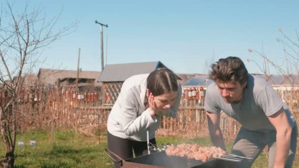 Pareja hombre y mujer cocinan carne shashlik en la parte superior de la parrilla de carbón en el patio trasero. Hablando y sonriendo juntos . — Vídeo de stock