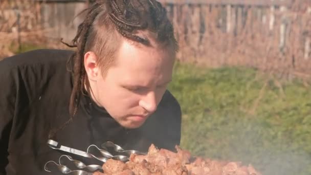 Jonge mannen met dreadlocks op haren koks sjasliek vlees bovenop de houtskool barbecue op achtertuin. — Stockvideo