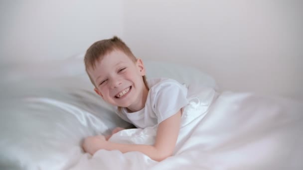 7 letni chłopiec tylko obudził się i śmieje się leżąc w łóżku. — Wideo stockowe