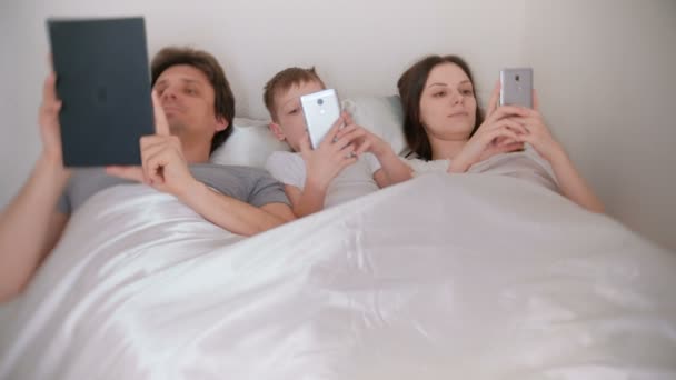 La familia está utilizando teléfonos móviles y tabletas que yacen en la cama por la mañana . — Vídeos de Stock