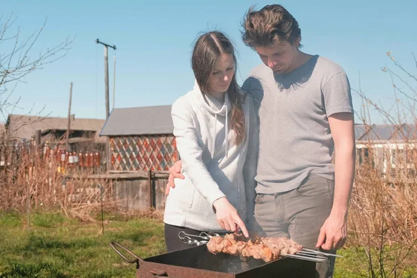 Casal homem e mulher cozinham carne shashlik em cima da grelha de carvão no quintal. Conversando e sorrindo juntos . — Fotografia de Stock