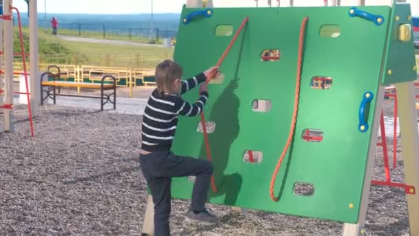 Хлопчик піднімається на скелелазну стіну, тримаючи мотузку на дитячому майданчику . — стокове відео
