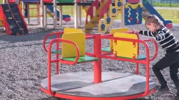 Niño de siete años girando en el carrusel en el patio de recreo . — Vídeo de stock