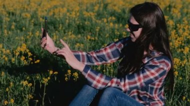 Kadın telefonda sarı çiçekler arasında çimlerin üzerine oturan bir selfie yapar.