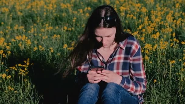 Donna che guarda il cellulare seduto nel parco sull'erba tra i fiori gialli . — Video Stock