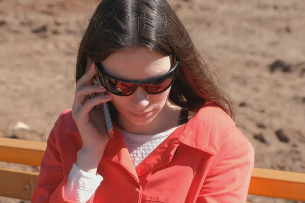 Ung brunett flicka solglasögon som väntar på ett samtal på telefonen sitter i parken på bänken. — Stockfoto