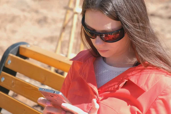 Joven morena en gafas de sol mira un video en el teléfono sentado en el parque en el banco . — Foto de Stock
