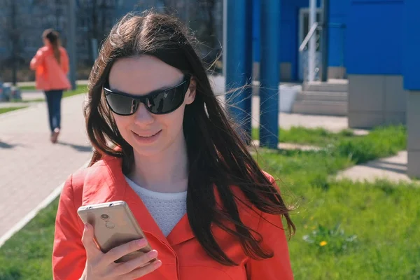 Jovem morena em óculos de sol em casaco vermelho espera por alguém e verifica seu telefone, mensagens de texto . — Fotografia de Stock