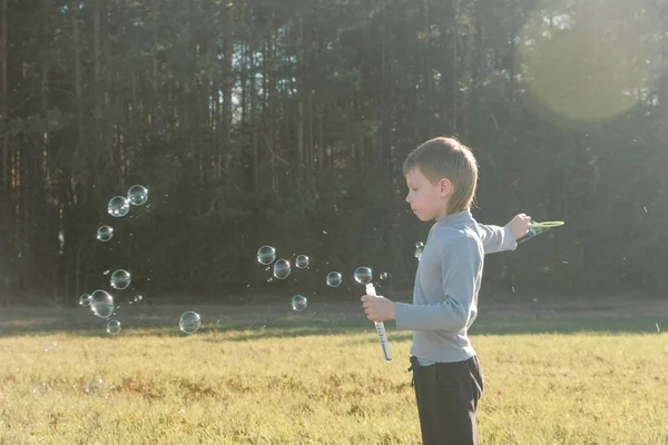 Pojken gör såpbubblor i en äng och spelar på en varm vårdag. — Stockfoto