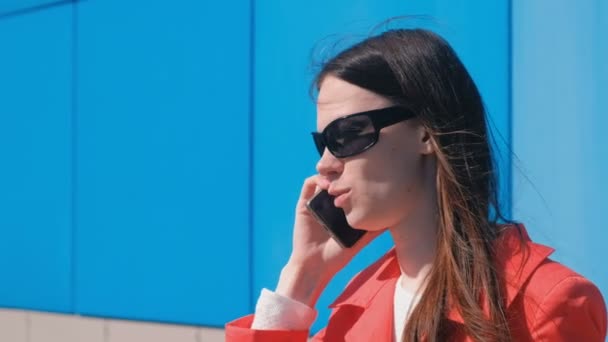 Retrato de jovem morena em óculos de sol fala ao telefone ao lado do prédio azul . — Vídeo de Stock