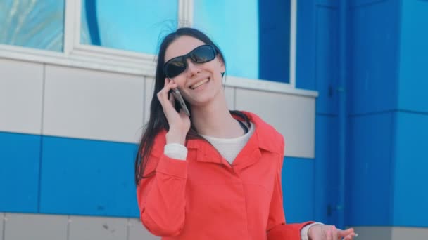 Porträtt av glad ung brunett kvinna i solglasögon talar i telefon den bredvid blå byggnad. — Stockvideo