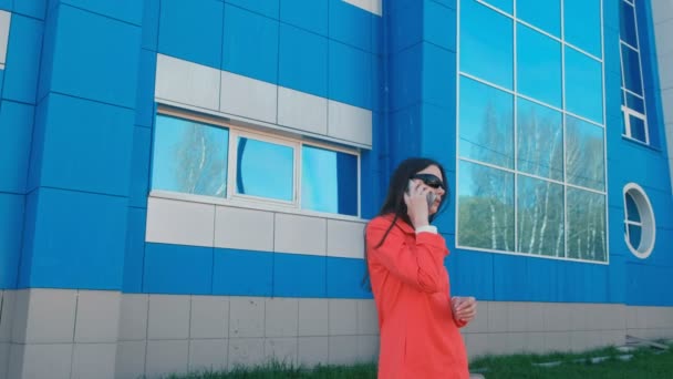 Portré, a boldog fiatal, barna nő napszemüveg beszél a telefonon a kék épület mellett. — Stock videók
