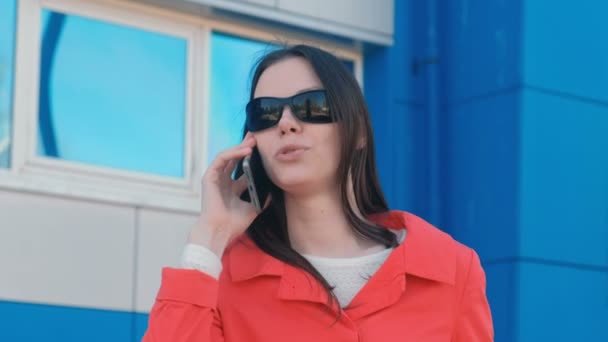Portré, fiatal, barna nő, napszemüveg és a piros kabát beszél a telefon mellett kék épület. — Stock videók