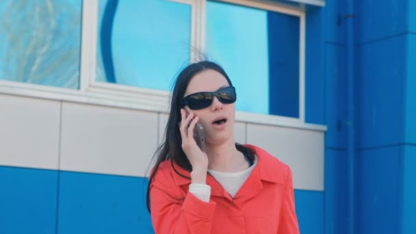 Portret van jonge brunette vrouw in zonnebrillen en rode jas spreekt op de telefoon naast blauwe gebouw. — Stockvideo