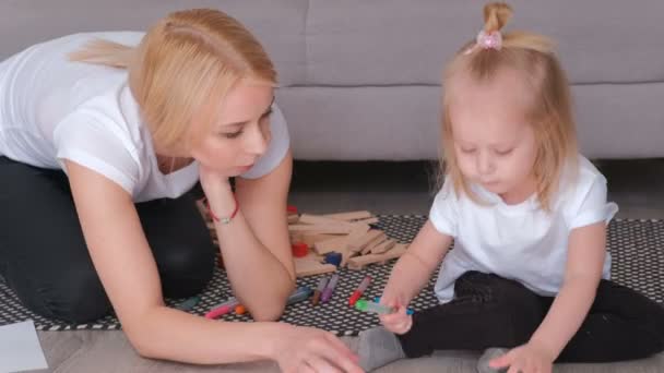 Kleines charmantes blondes Mädchen bemalt Filzstift mit ihrer schönen Mutter, die neben dem Sofa sitzt. Nahaufnahme. — Stockvideo