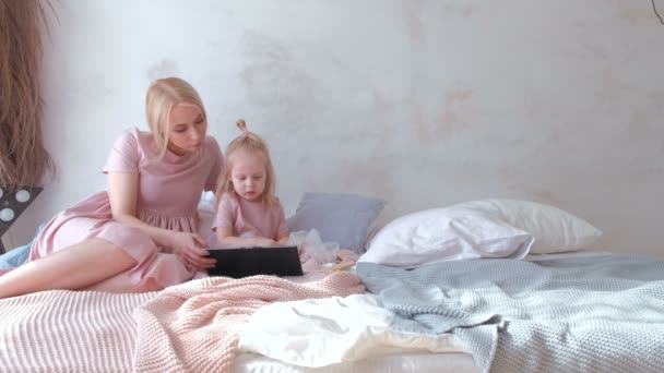 Joven mujer rubia atractiva con su pequeña hija encantadora en vestidos de color rosa viendo algo en la tableta y hablando . — Vídeos de Stock