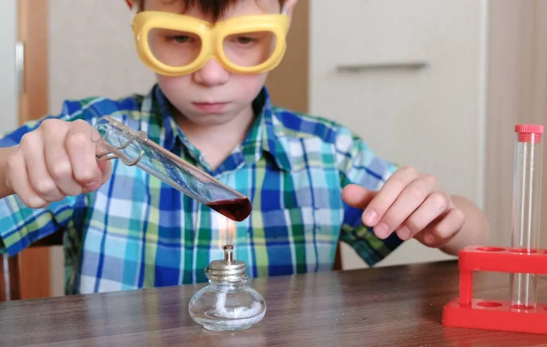 Experimentos de química en casa. Chico calienta el tubo de ensayo con líquido rojo en la lámpara de alcohol ardiente . — Foto de Stock