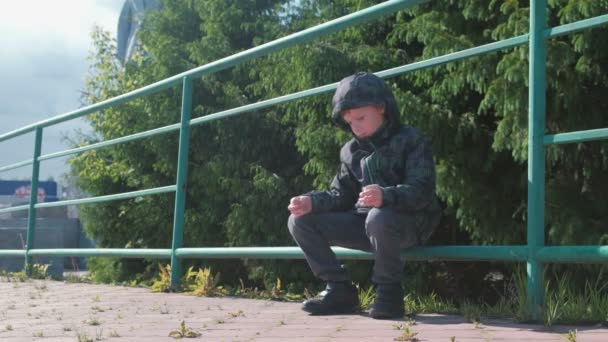 Niño de siete años sentado en una valla en el parque y mira a su alrededor . — Vídeos de Stock