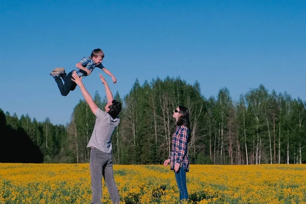 Papá lanza a su hijo al aire, y la madre sigue. Paseo familiar por el campo con flores amarillas cerca del bosque . — Foto de Stock