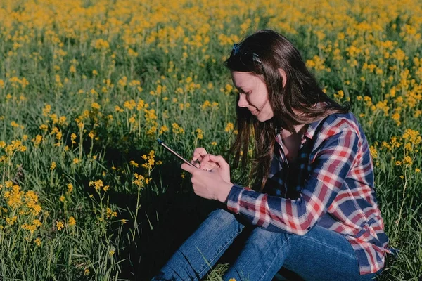 Mulher olhando para o telefone móvel sentado no parque na grama entre flores amarelas . — Fotografia de Stock